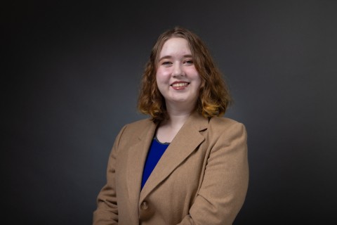 White woman with brown shoulder length hair. She is wearing a tan blazer over a blue blouse. 